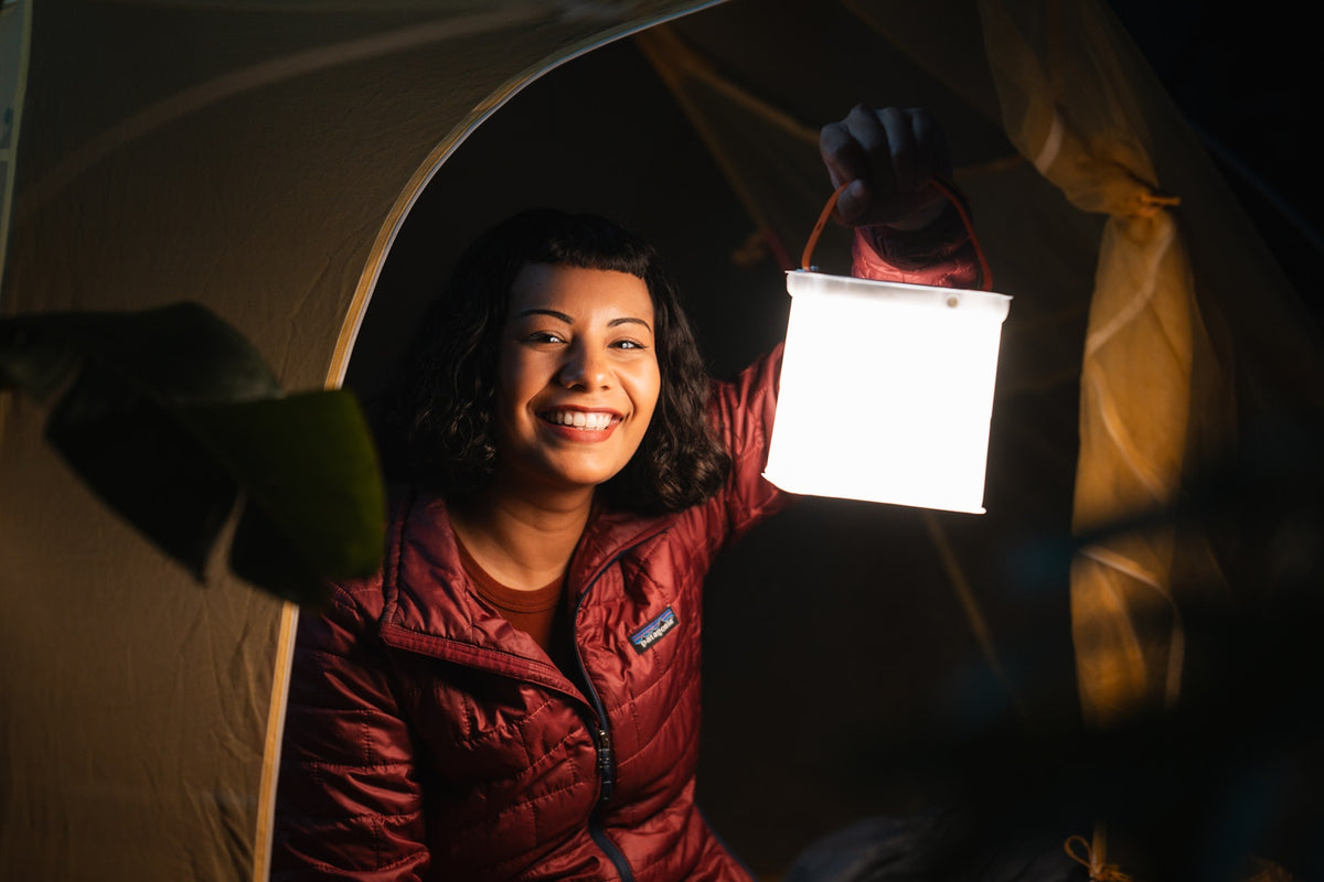 A smiling woman inside a tent at night holds up a glowing white lantern. She is wearing a dark red puffer jacket with a visible Patagonia logo on the chest.