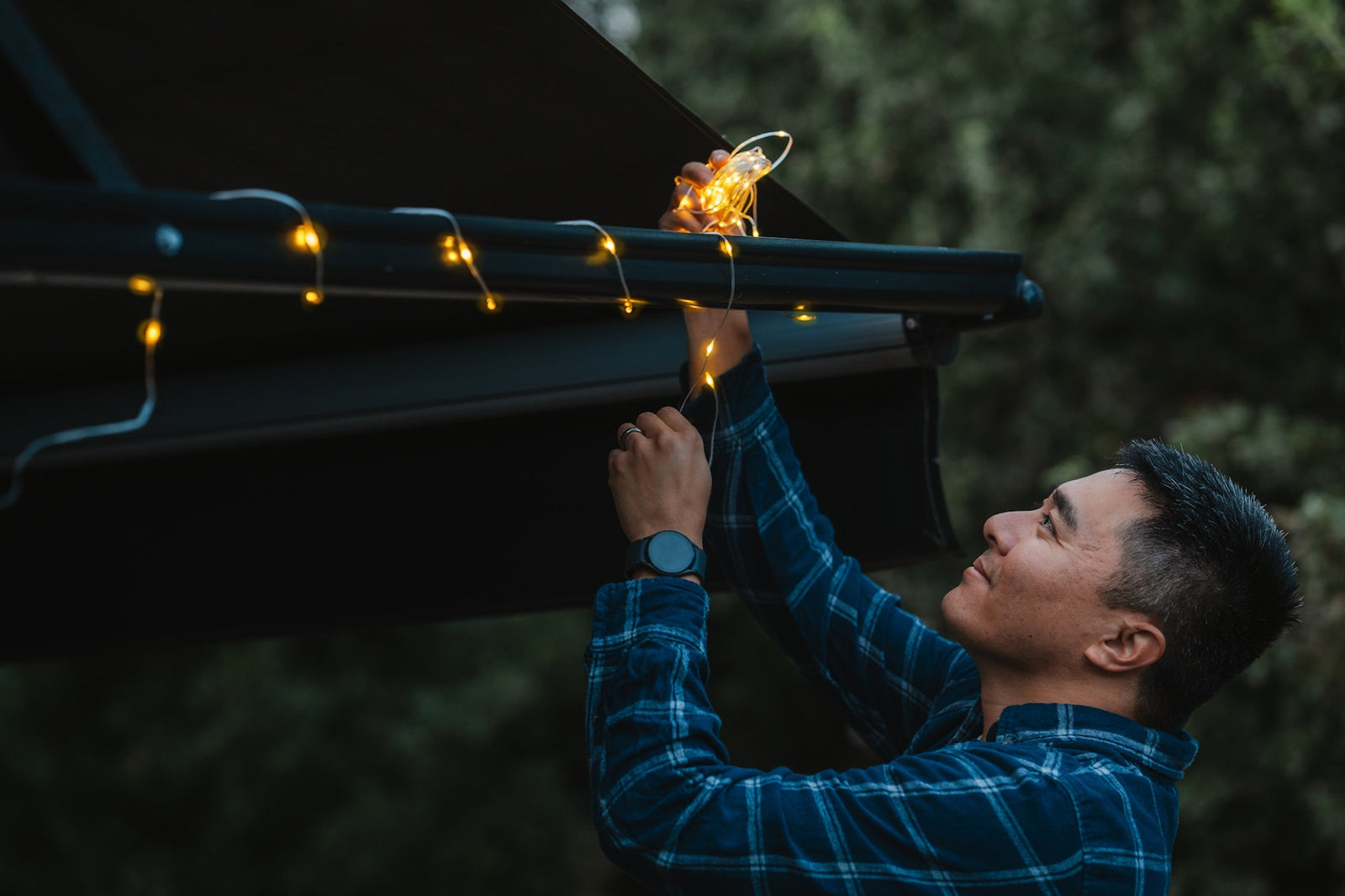 A man in a blue plaid shirt hangs warm white string lights along the edge of a black piano outdoors at dusk.