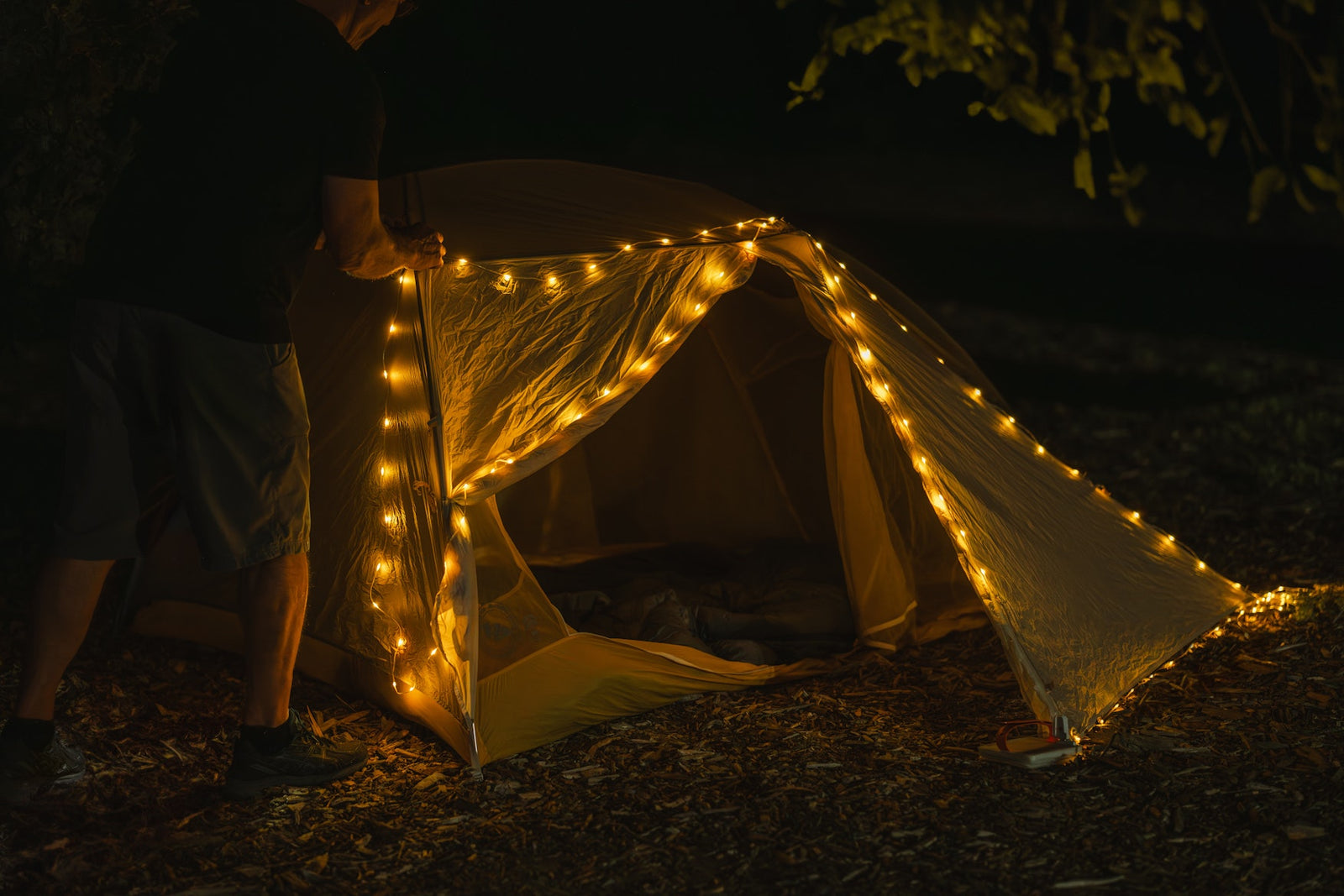 A person at night sets up a dome camping tent outdoors, with warm string lights glowing around the tent entrance and along the edges.