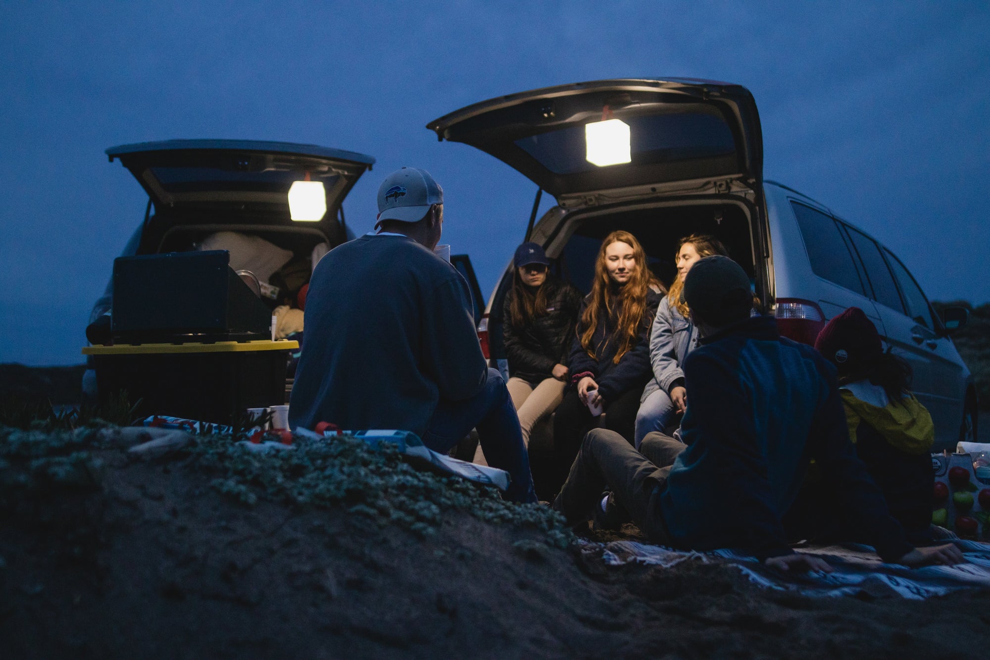 group of people sitting outside of two vehicles with lanterns hanging off of the open trunk of the vehicle