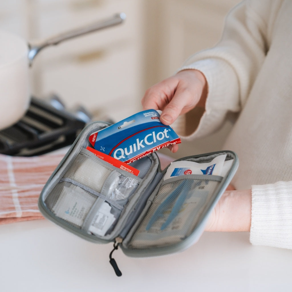 A gray zippered first aid kit held open by a person in a cream sweater, showing medical supplies including a blue and red QuikClot pack inside mesh compartments.
