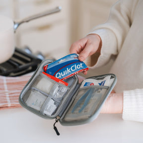 A gray zippered first aid kit held open by a person in a cream sweater, showing medical supplies including a blue and red QuikClot pack inside mesh compartments.