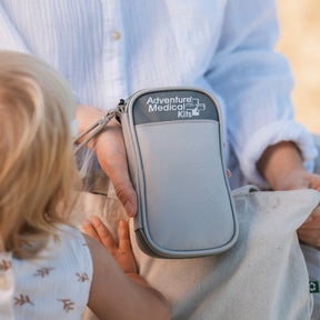 A light gray Adventure Medical Kits zippered pouch held by an adult outdoors while a child sits beside them. The pouch has a dark gray top label with white text and a wrist strap.