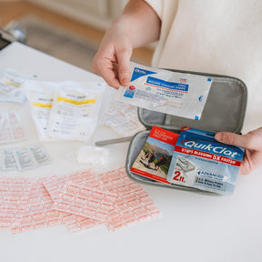 A person holds an open gray mesh first-aid kit on a white table, showing QuikClot blood pressure dressing packets and other medical supplies. Several wrapped bandages are spread out on the table.
