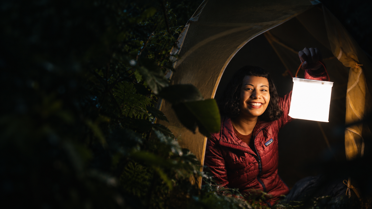 A smiling woman in a red jacket sits inside a camping tent at night, holding up a glowing lantern. Dark foliage frames the left side of the scene.